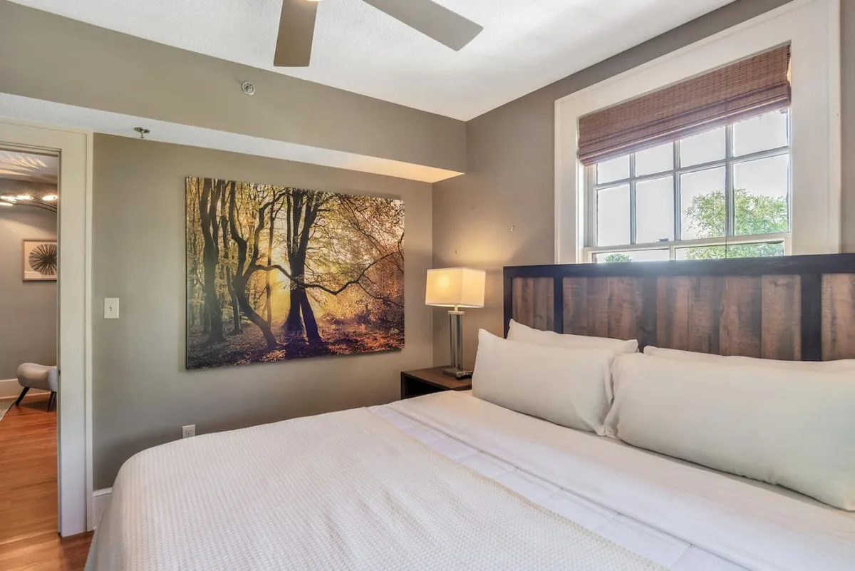 A serene king bedroom with white linens, calming artwork of trees above the bed, a lamp on the nightstand, and ample natural light flooding through the window. The atmosphere is peaceful, perfect for restful sleep or a quiet moment with a book.