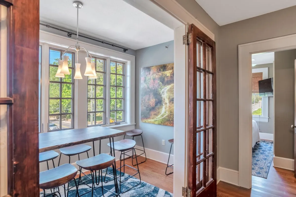 The image features a sunlit interior space with large windows that allow for urban views, a wooden table with stools, and artistic decor on the wall. The space feels open and welcoming, capturing the airy, elevated feel of an upper-floor city apartment. The ample natural light and urban backdrop through the windows evoke the sense of being above the city bustle.