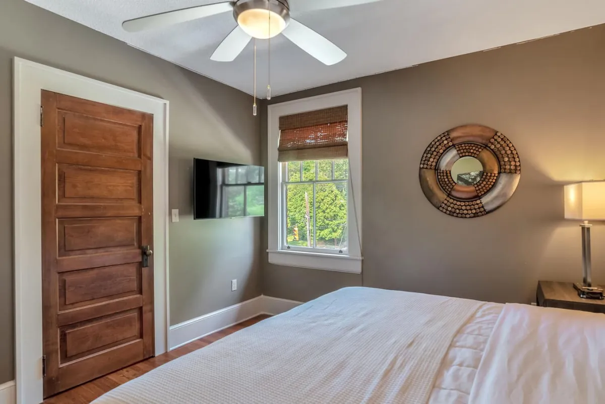 A cozy bedroom featuring a plush bed, wall mirror, window, and TV. The room feels serene and private, ideal for unwinding, reading, or enjoying a quiet evening in after exploring downtown. The view through the window reinforces a connection to the outside world.