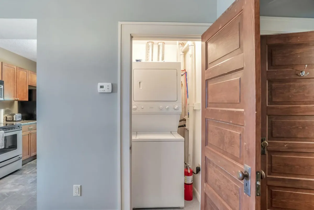 The image captures a practical corner of the home with a washer and dryer set neatly into a nook, with a glimpse of the kitchen area visible in the background. The scene emphasizes home convenience and the seamless integration of functional spaces. Warm lighting and tidy design contribute to a welcoming, organized environment.