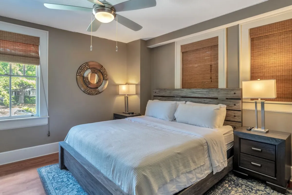 A tranquil king bedroom featuring a bed with crisp white linens, a wooden headboard flanked by nightstands each with a lamp, a decorative wall mirror, and a window streaming in natural light. The space feels inviting and cozy, ideal for unwinding after a day in Asheville.